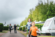 Two children, a boy and a girl, are running past touring vans next to trees and two adults walk in the background 