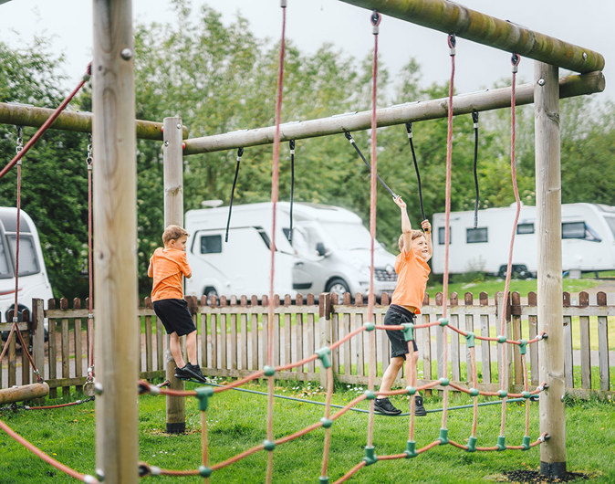 Twin boys in matching shirts and a young girl playing and climbing on rope apparatus with touring vans in the background